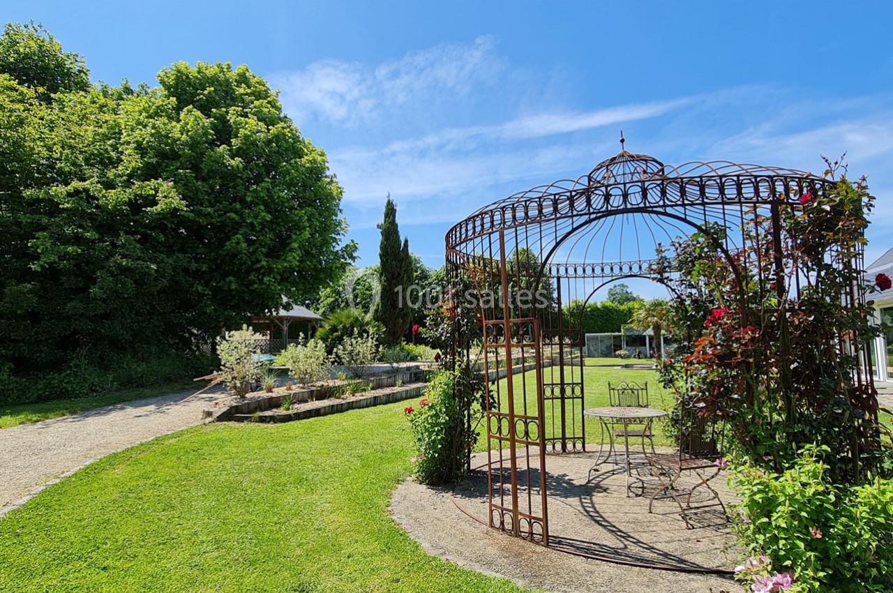 Tonnelle en fer forgé entourée de rosiers dans un jardin verdoyant, avec un chemin et des arbres en arrière-plan.