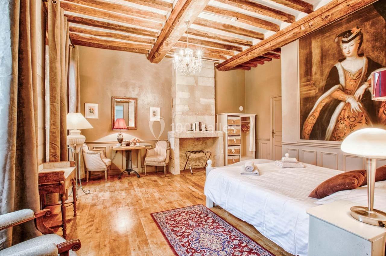 Chambre lumineuse avec poutres apparentes, lit double, coin bureau, cheminée en pierre et grand tableau mural.