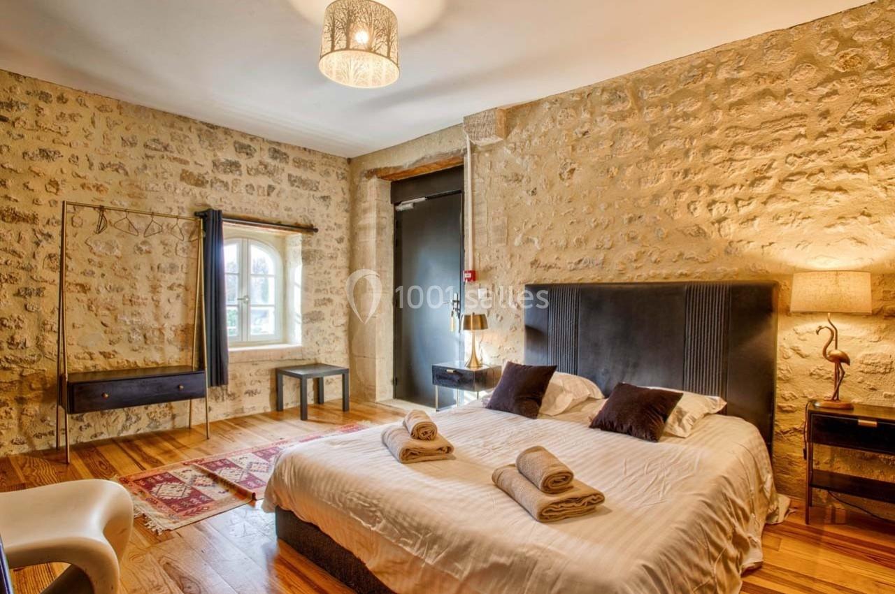 Chambre avec murs en pierre, lit double, lampes, fenêtre lumineuse et décoration chaleureuse sur parquet en bois.