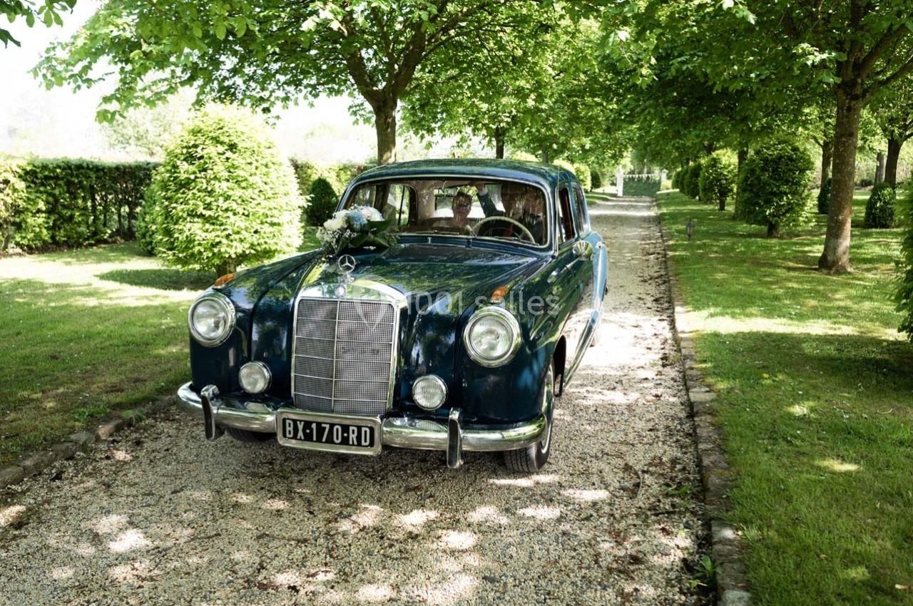 Voiture ancienne bleue circulant sur une allée gravillonnée bordée d’arbres et de verdure.