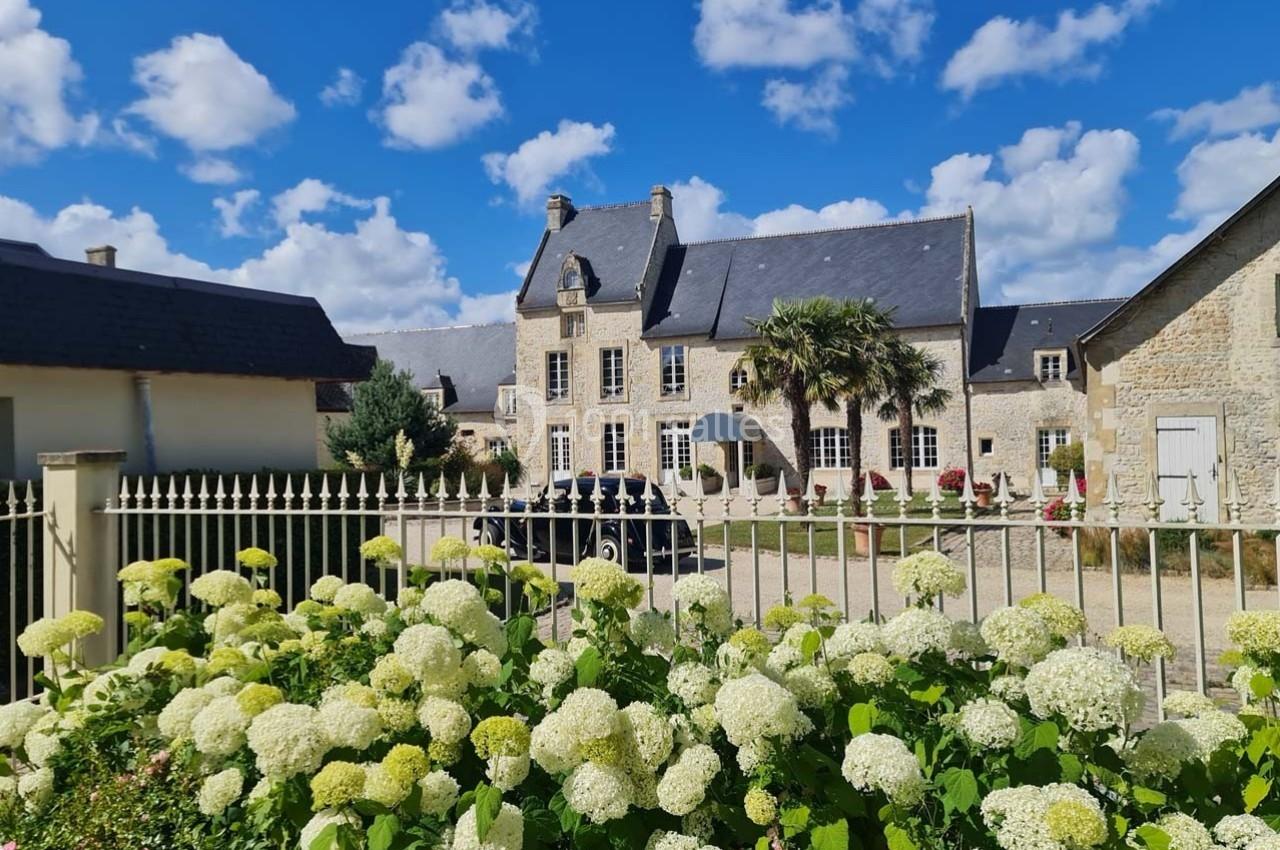 Façade d'une maison en pierre avec toit en ardoise, jardin fleuri au premier plan et ciel bleu parsemé de nuages.