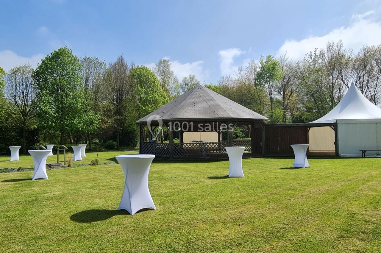 Espace extérieur avec pelouse, tables hautes couvertes de nappes blanches, kiosque en bois et tente blanche.