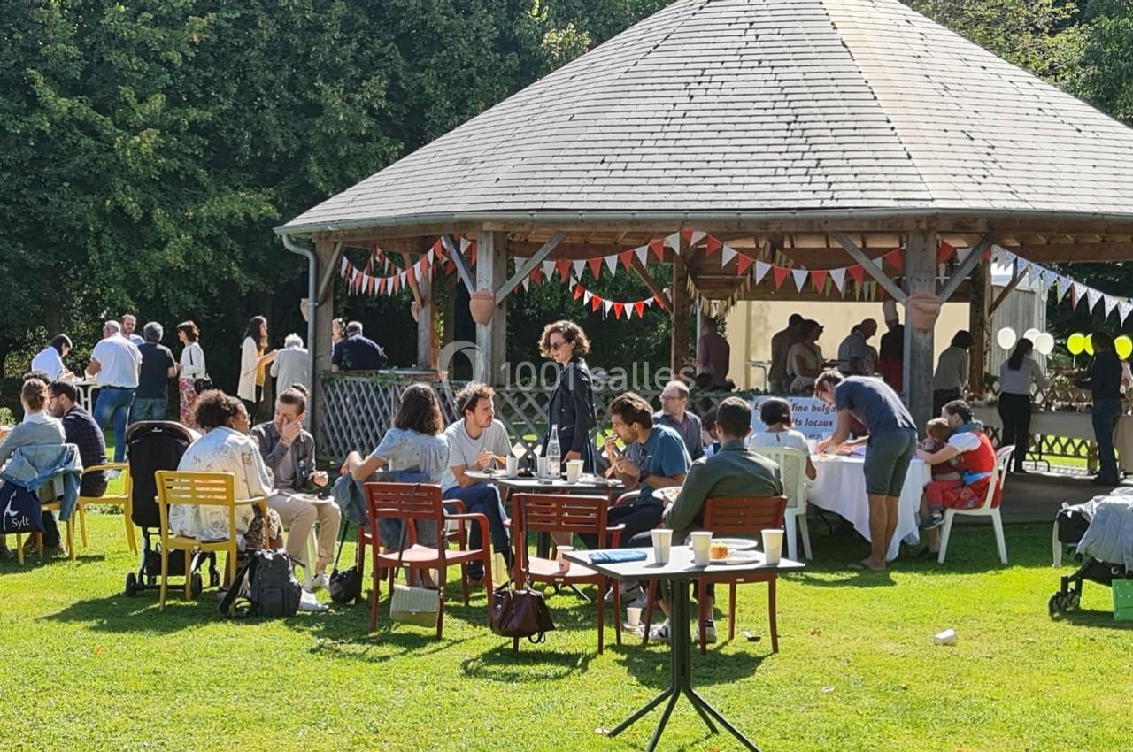 Des personnes assises et debout discutent autour de tables dans un jardin, près d'un kiosque décoré de fanions.