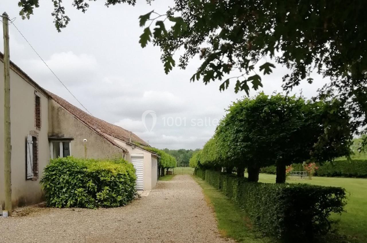 Allée gravillonnée bordée de haies taillées, menant à une maison en pierre sous un ciel nuageux.