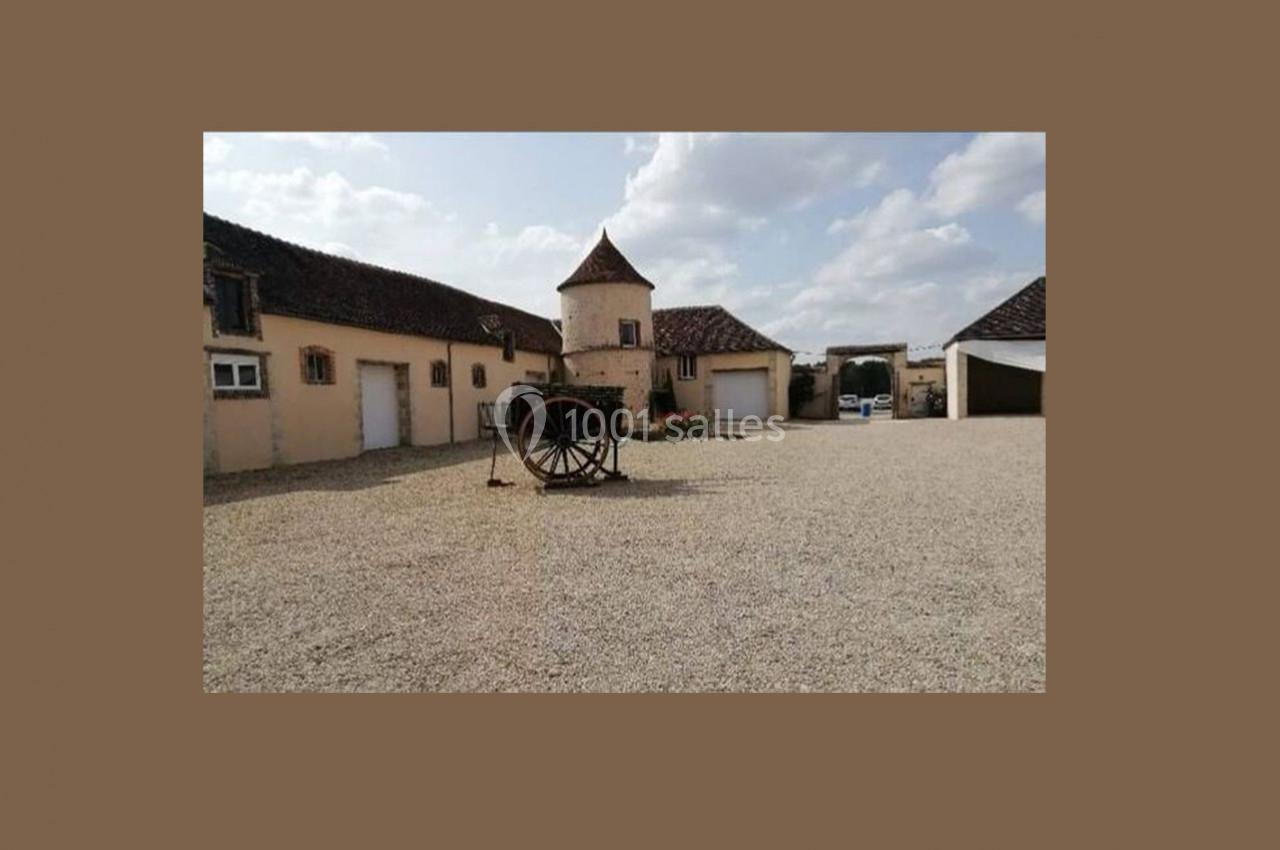 Cour en gravier avec bâtiments en pierre, une charrette en bois au centre et un ciel partiellement nuageux.