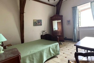 Chambre avec poutres apparentes, lit simple, armoire ancienne, bureau, chaise et fenêtre ouverte sur l'extérieur.