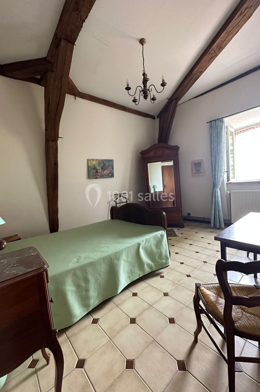 Chambre avec poutres apparentes, lit simple, armoire ancienne, bureau, chaise et fenêtre ouverte sur l'extérieur.