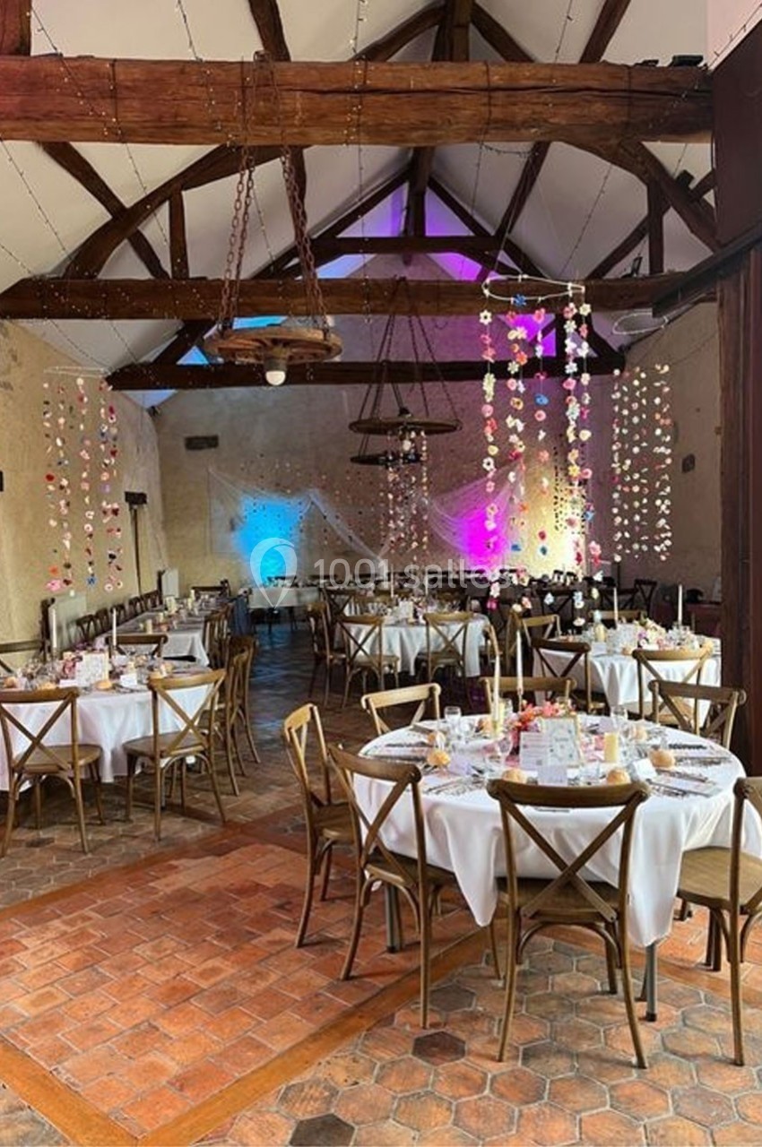 Salle de réception décorée avec des tables rondes dressées, guirlandes suspendues et éclairage coloré.