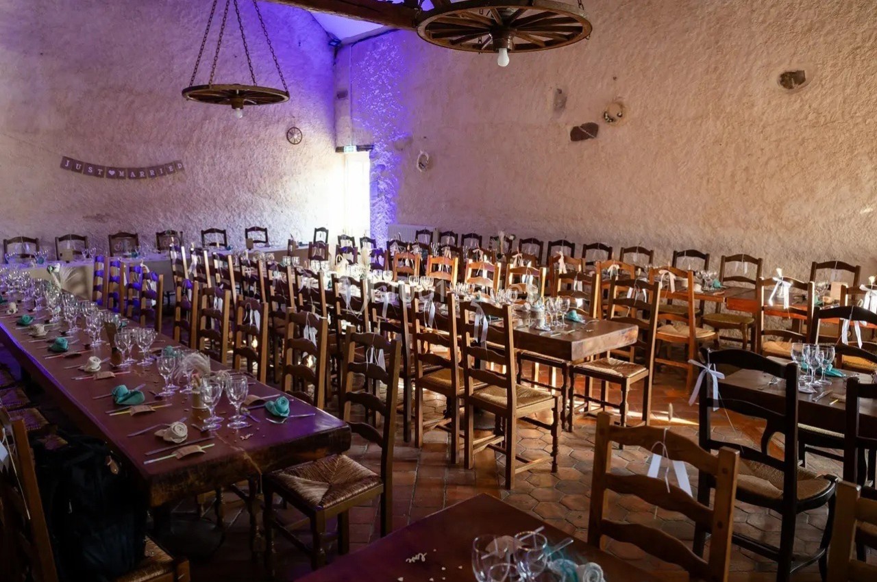 Salle de réception rustique avec de longues tables en bois dressées, entourées de chaises, éclairée par une lumière douce.