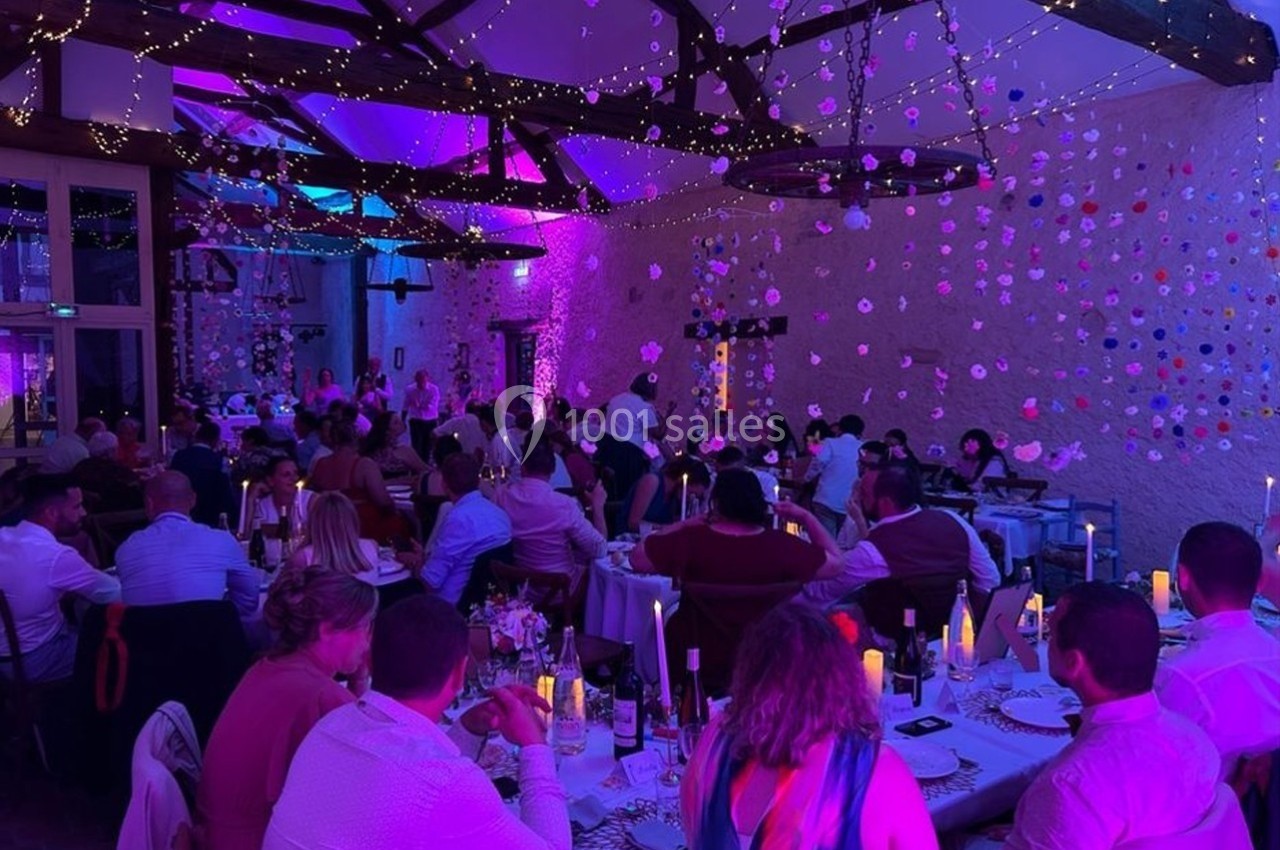 Salle de réception décorée avec guirlandes lumineuses et fleurs suspendues, des invités dînent à des tables rondes.