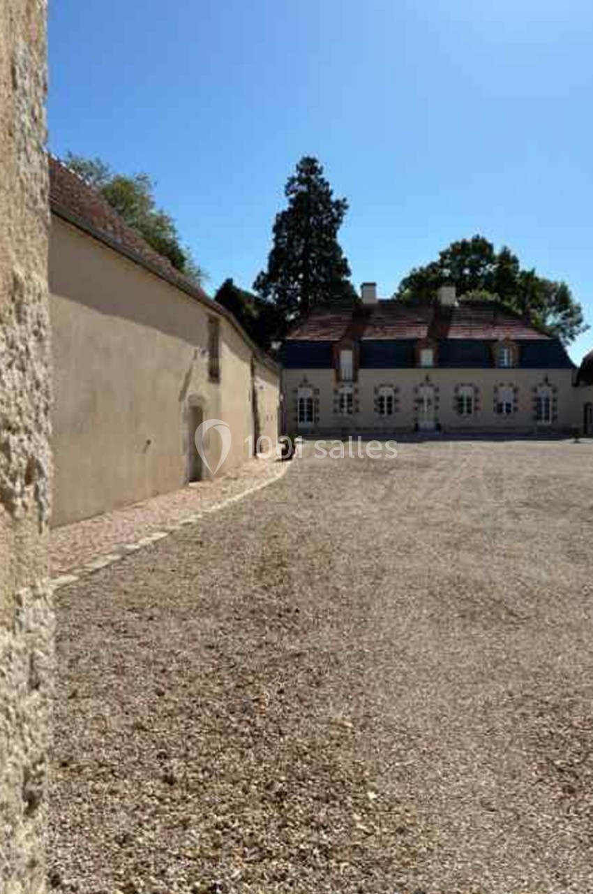 Cour en gravier bordée de bâtiments anciens avec toits en tuiles, sous un ciel dégagé.