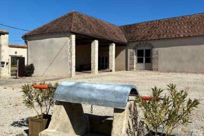 Cour pavée avec un puits en pierre au premier plan, entouré de bacs végétalisés, et bâtiments en arrière-plan.