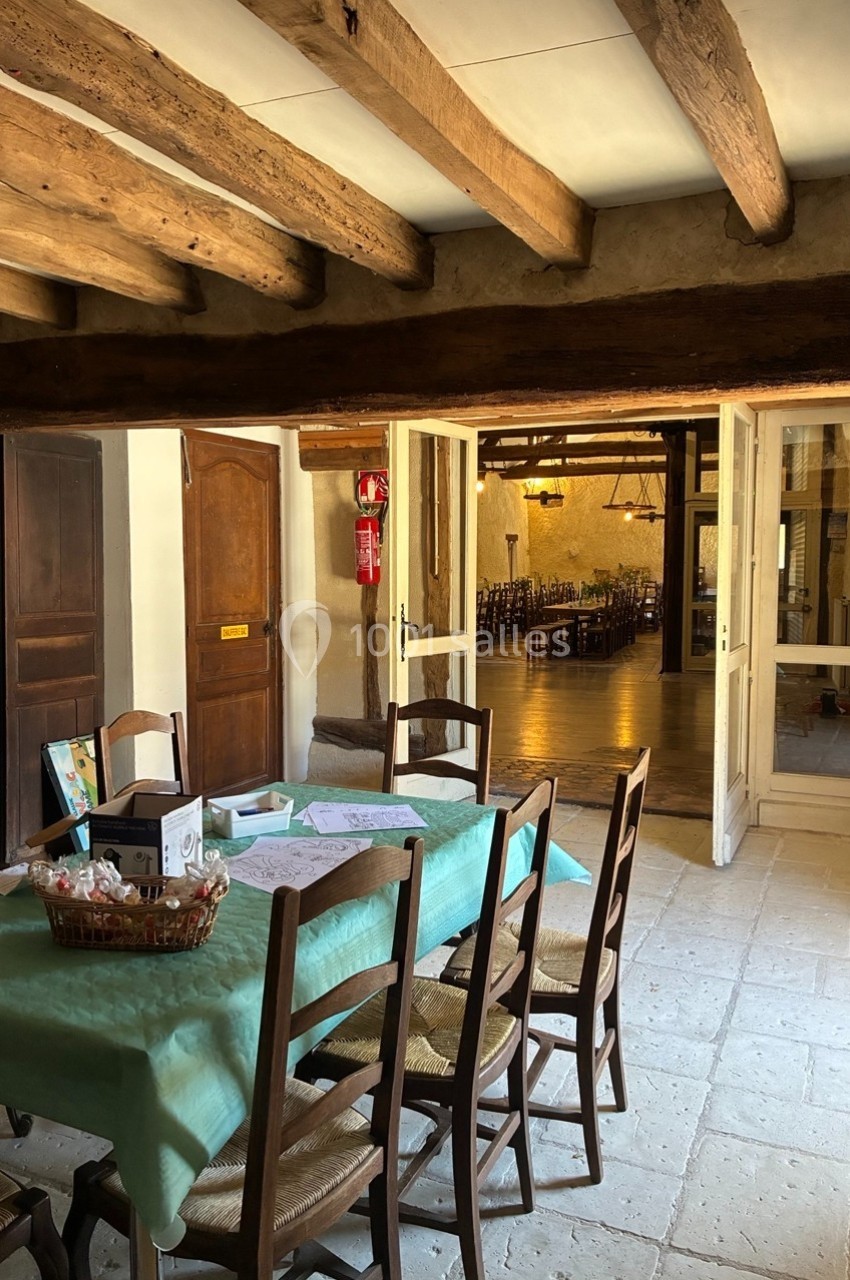 Salle à manger rustique avec poutres apparentes, table dressée et vue sur une grande pièce éclairée en arrière-plan.