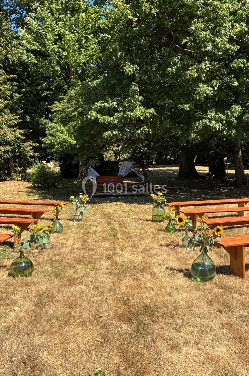 Allée centrale bordée de bancs en bois et de vases avec des tournesols, menant à une arche décorée dans un jardin ombragé.