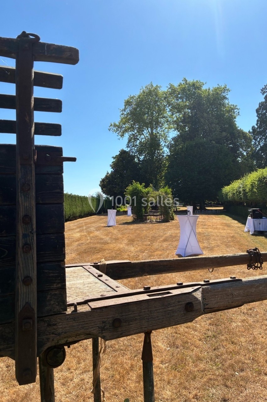 Vue d'un jardin ensoleillé avec des tables hautes drapées de blanc, entouré de verdure et d'arbres.