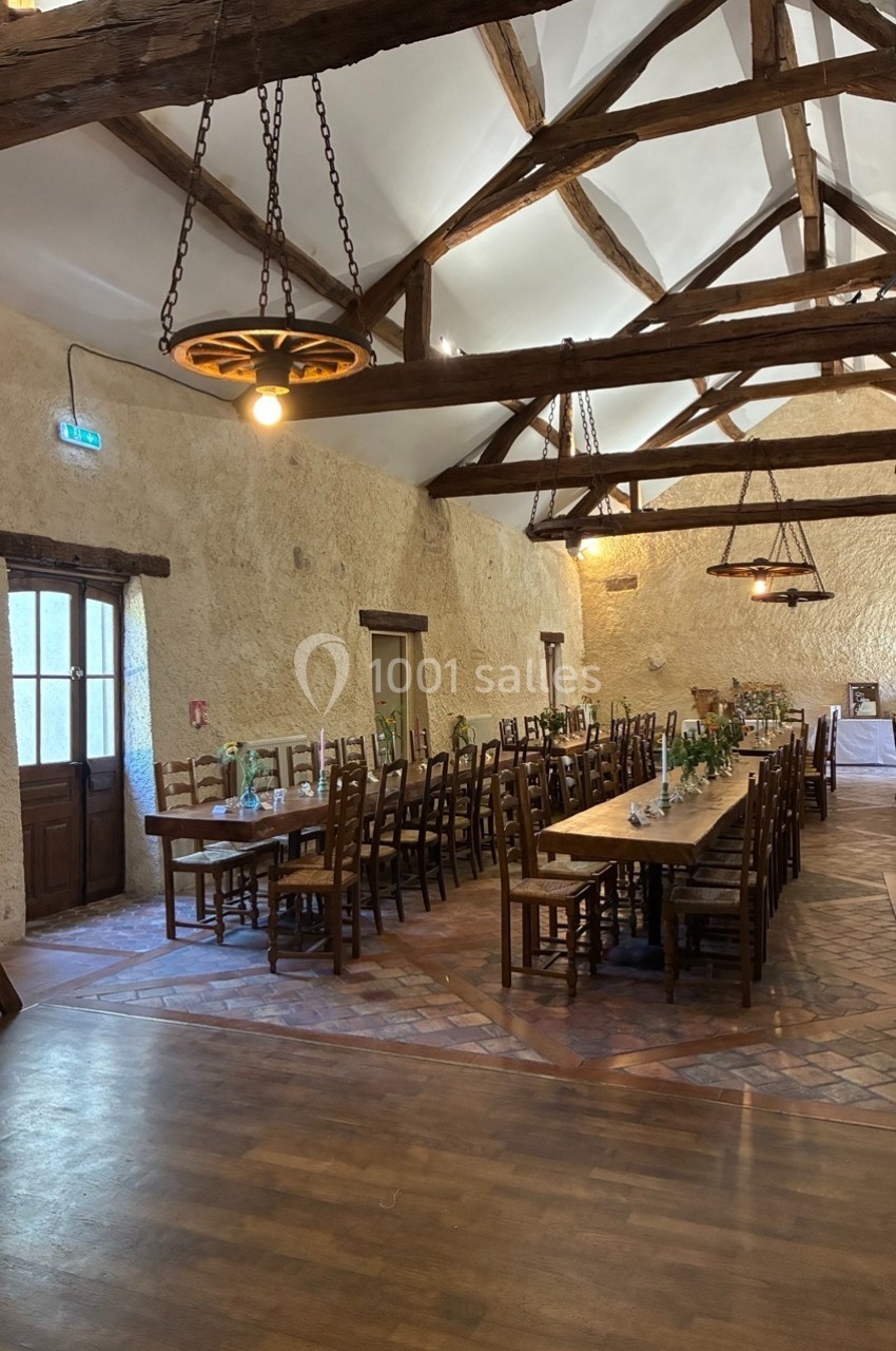 Salle rustique avec poutres apparentes, tables en bois alignées et décorations florales, éclairée par des suspensions.