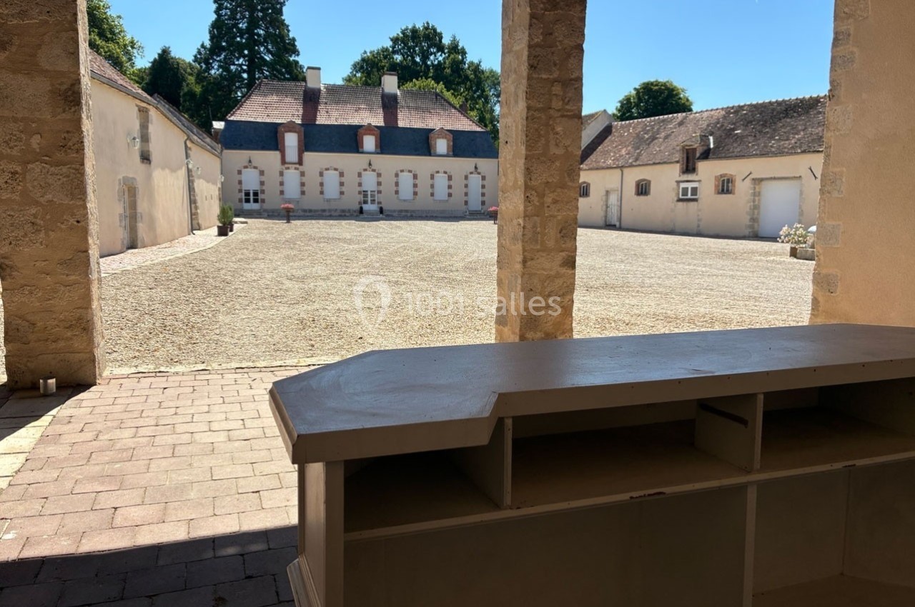 Vue d'une cour pavée entourée de bâtiments anciens, prise depuis un porche avec un comptoir en bois au premier plan.