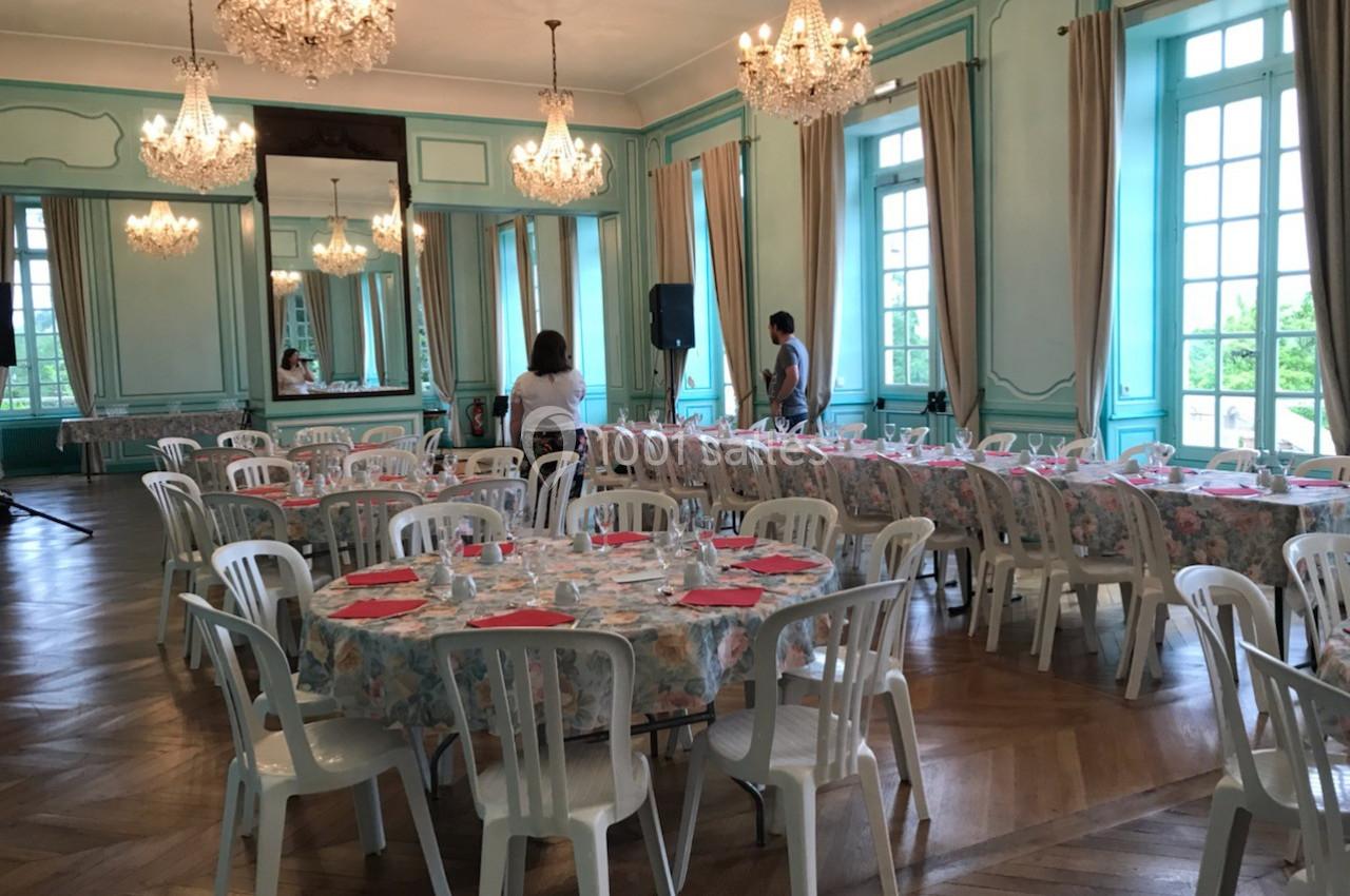 Salle de réception lumineuse avec tables rondes dressées, chaises blanches et lustres suspendus.