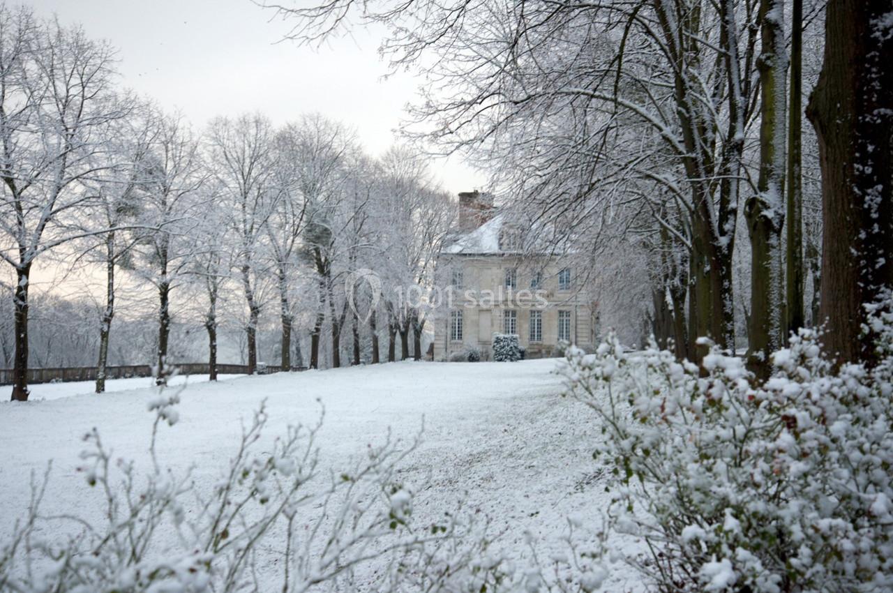 Manoir entouré d'arbres et d'un jardin recouverts de neige en hiver, sous un ciel nuageux.