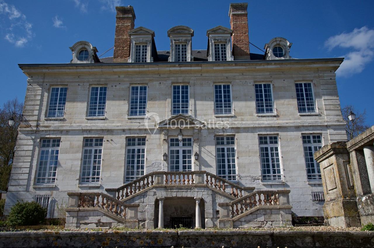 Façade d'un manoir en pierre avec escaliers symétriques, grandes fenêtres et cheminées, sous un ciel dégagé.