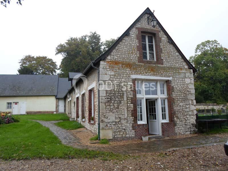 Bâtiment en pierre avec une grande porte vitrée, entouré de verdure et d'une allée pavée sous un ciel nuageux.