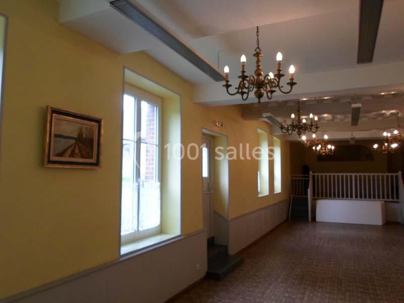 Salle lumineuse avec murs jaunes, lustres suspendus, tableau encadré et escalier menant à une mezzanine.