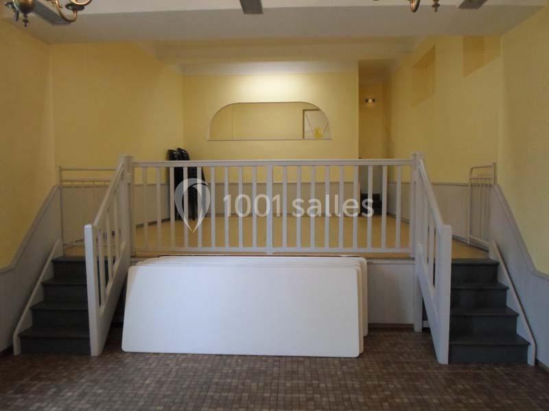 Salle vide avec murs jaunes, estrade surélevée accessible par deux escaliers et rambardes blanches.