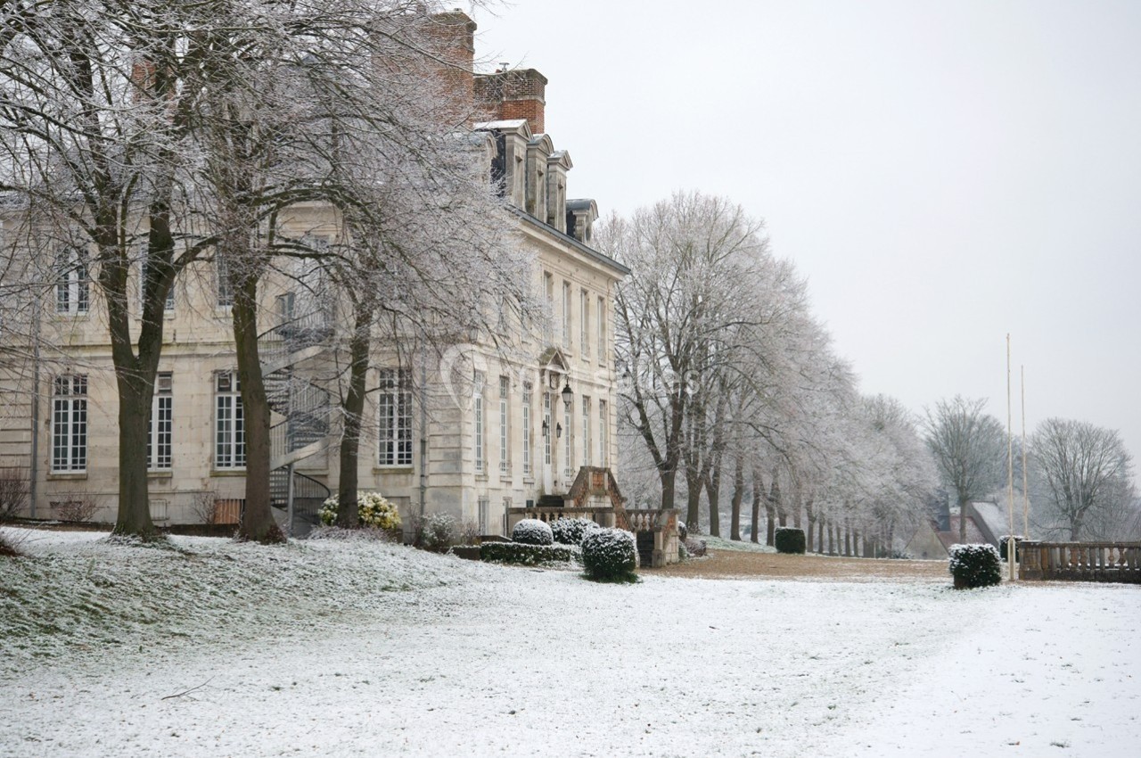 Façade d'un bâtiment ancien entouré d'arbres et d'un jardin recouvert de neige en hiver.