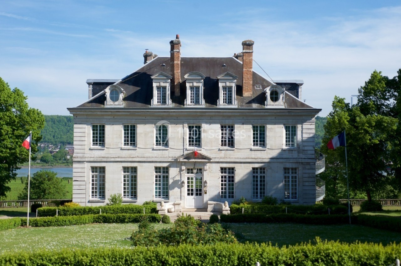Façade d'un manoir en pierre entouré d'un jardin avec drapeaux français, sous un ciel dégagé.