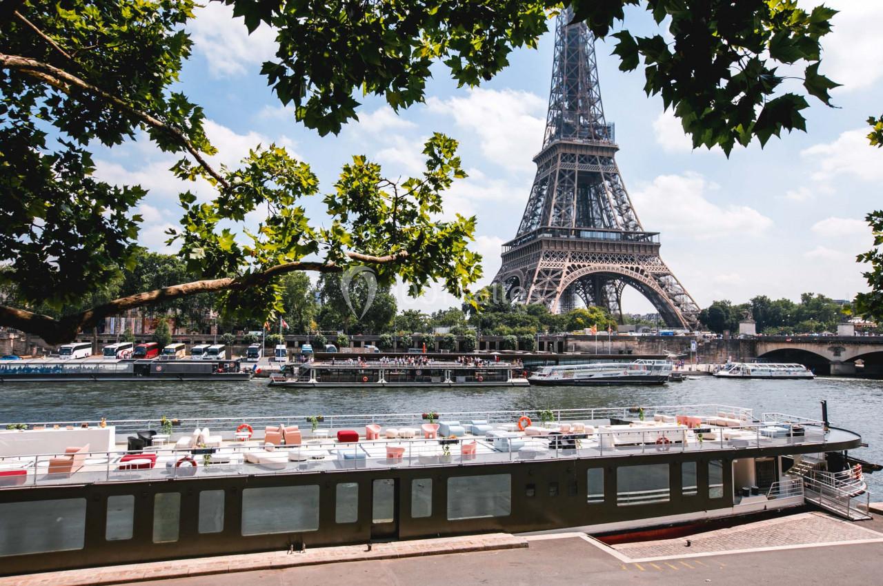 Vue de la Tour Eiffel depuis les quais de la Seine, avec des péniches amarrées et des arbres encadrant la scène.