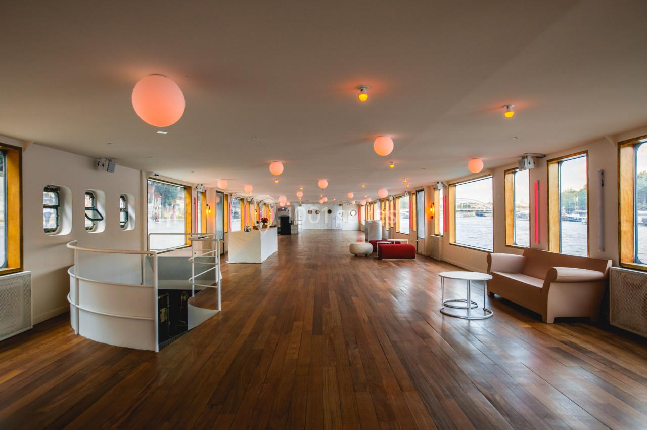 Salle spacieuse avec parquet en bois, canapés, baies vitrées et éclairage suspendu, offrant une vue sur l'extérieur.