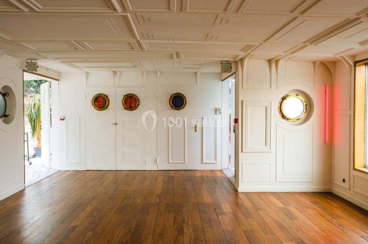 Salle lumineuse avec parquet en bois, murs blancs ornés de hublots et de moulures, et une lumière néon rose.
