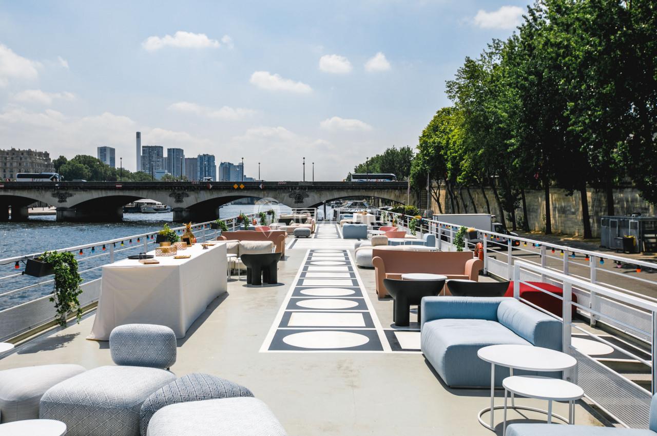 Terrasse aménagée d'un bateau avec mobilier moderne, vue sur un pont et des immeubles en arrière-plan.