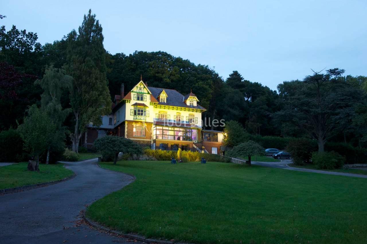 Grande maison éclairée au crépuscule, entourée d'un jardin verdoyant et bordée d'une allée sinueuse.