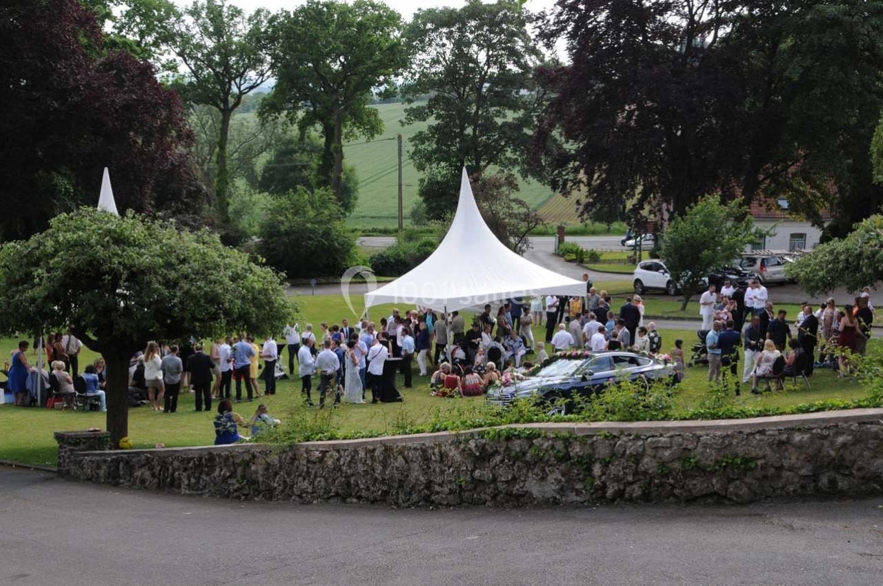 Groupe de personnes rassemblées sous une tente blanche dans un jardin verdoyant, avec des arbres et des voitures en arrière…
