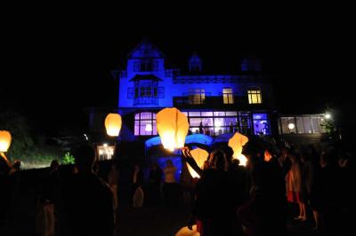 Des lanternes lumineuses flottent dans la nuit près d'une grande maison éclairée en bleu, entourée de personnes.