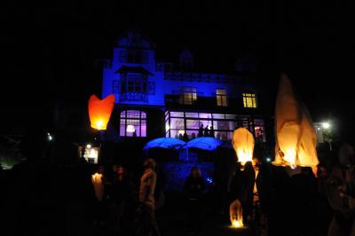 Des lanternes lumineuses flottent dans la nuit près d'une grande maison éclairée en bleu, entourée de personnes.
