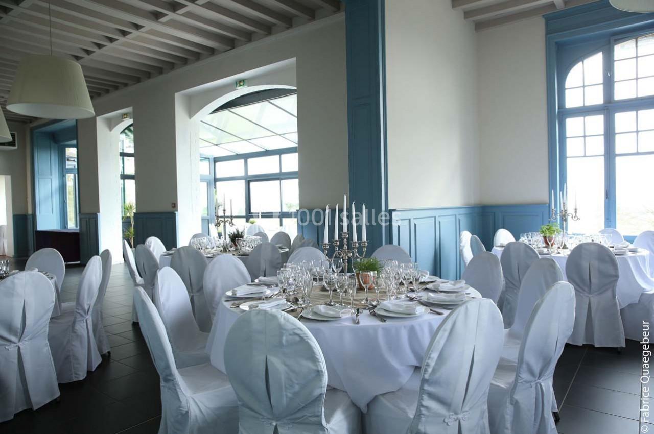 Salle de réception lumineuse avec tables rondes dressées, nappes blanches et chandeliers élégants.