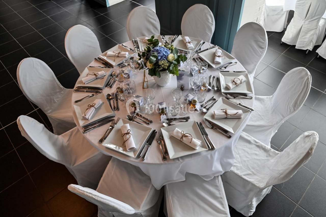 Table ronde dressée avec nappes blanches, couverts, serviettes pliées et centre de table floral, entourée de chaises…