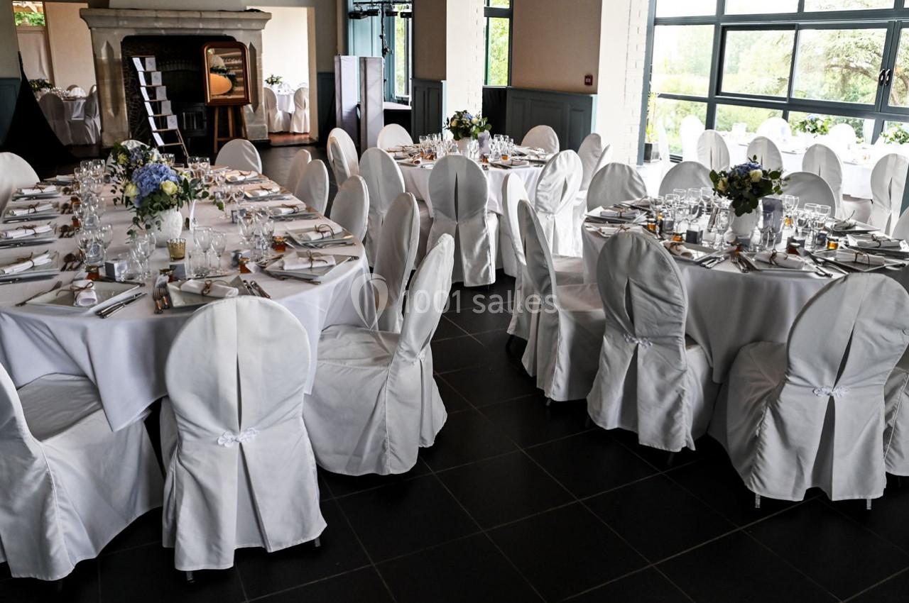 Salle de réception avec tables rondes dressées, nappes blanches, chaises houssées et décorations florales.