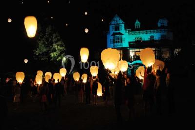Des lanternes lumineuses flottent dans la nuit près d'une grande maison éclairée en bleu, entourée de personnes.
