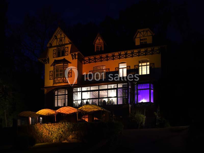 Façade d'une grande maison éclairée la nuit, avec des fenêtres illuminées et un jardin à l'avant.