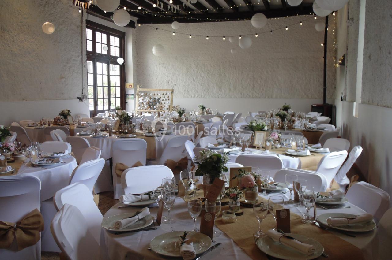 Salle décorée pour un mariage avec des tables rondes dressées, nappes blanches, chemins de table beige et guirlandes…