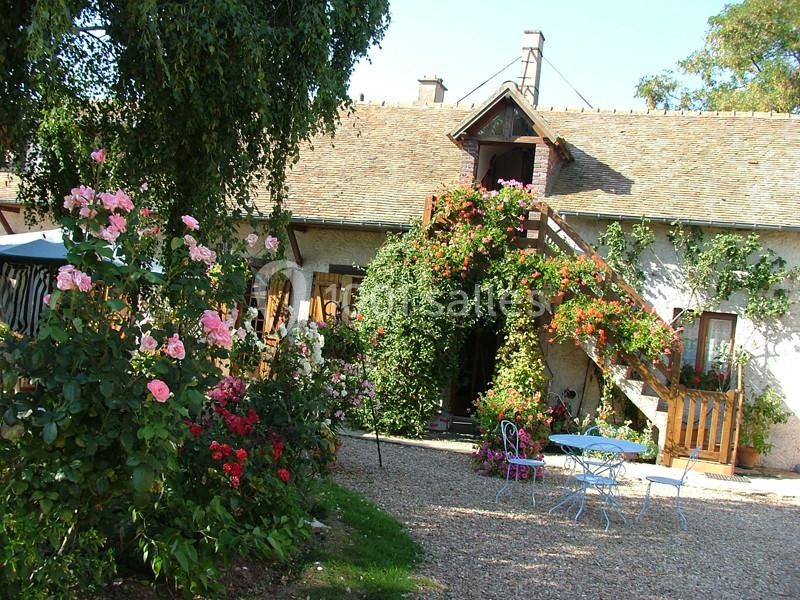 Maison de campagne avec escalier extérieur fleuri, jardin coloré et mobilier de jardin sur une allée gravillonnée.