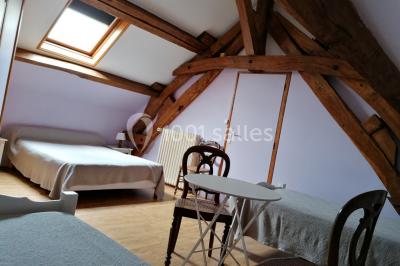 Chambre mansardée avec poutres apparentes, trois lits, une table et des chaises, éclairée par une fenêtre de toit.