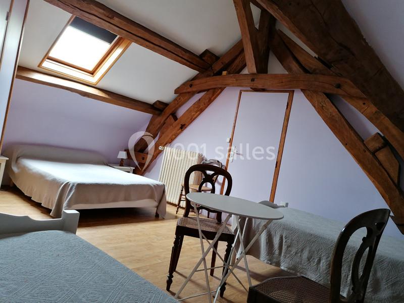 Chambre mansardée avec poutres apparentes, trois lits, une table et des chaises, éclairée par une fenêtre de toit.