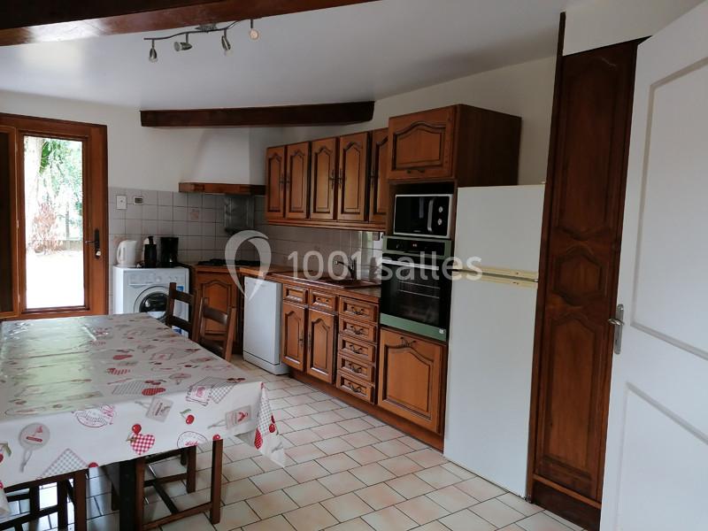 Cuisine équipée avec meubles en bois, table avec nappe, électroménagers et fenêtre donnant sur l'extérieur.
