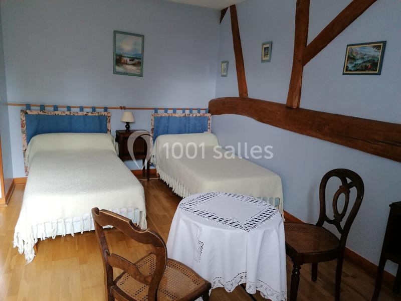 Deux lits simples dans une chambre aux murs bleus, avec poutres apparentes, meubles en bois et table ronde avec nappe.