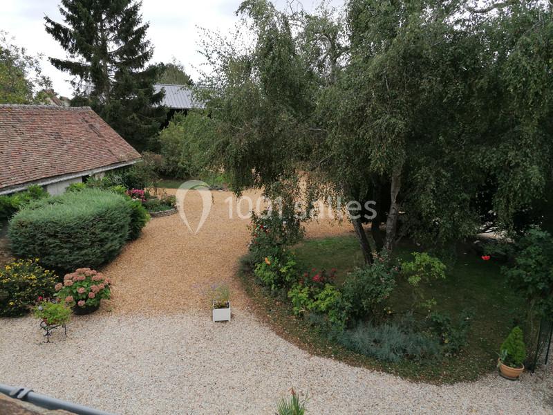 Vue d'une cour avec allée en gravier, arbres, arbustes fleuris et bâtiments en arrière-plan.