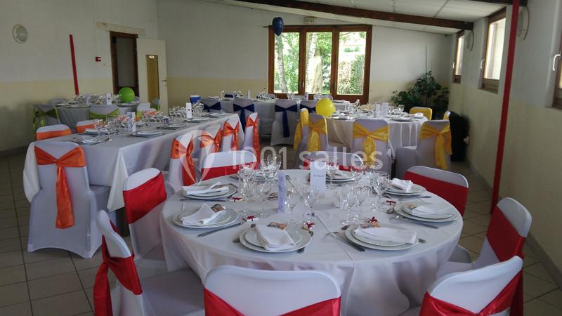 Salle décorée pour un événement avec des tables rondes dressées, nappes blanches et chaises ornées de nœuds colorés.